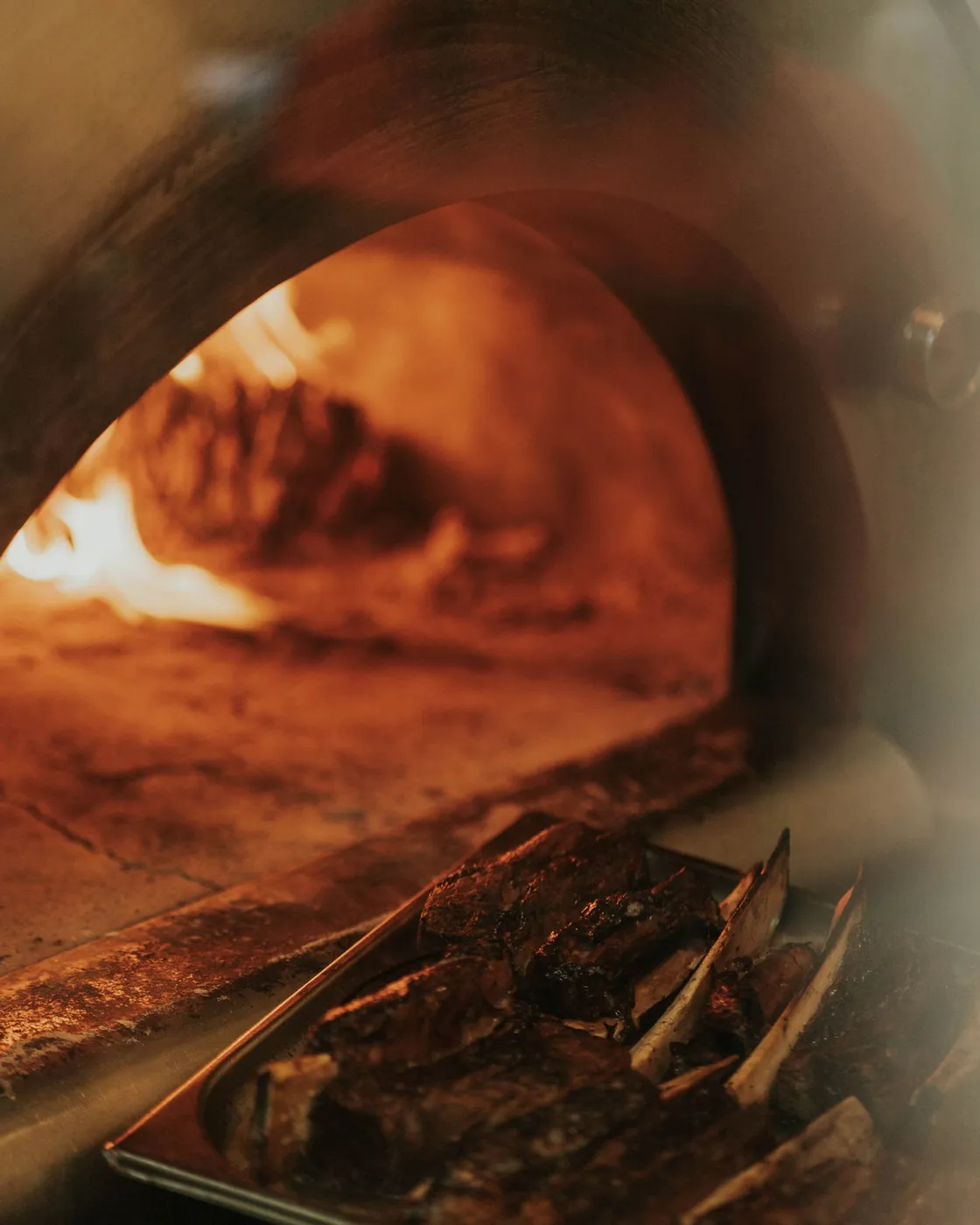Horno de leña encendido con una bandeja de carne asándose a la brasa